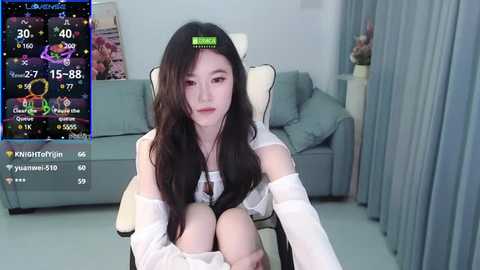 Media: A young Asian woman with long, dark hair sits on a white chair in a modern living room, wearing a white blouse. A digital screen displays a live stream overlay.