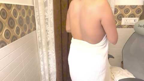 Media: Video of a nude woman, viewed from behind, wrapping a white towel around her body, standing in a bathroom with beige and brown patterned walls, a shower curtain, and a chair.