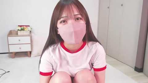 Media: Video of an Asian woman with long brown hair, wearing a white T-shirt and pink surgical mask, sitting on a white floor. Background features a white wall, dresser, and potted flowers.