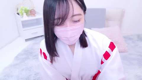 Media: Video of an Asian woman with shoulder-length black hair, wearing a white yukata with red accents and a pink face mask, kneeling on a light grey carpet in a minimalist, white room with a nightstand and potted plants.