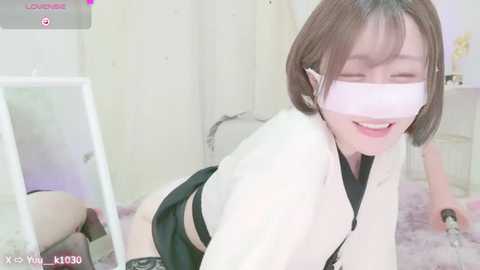 Media: Video of a petite Asian woman with short brown hair, wearing a white mask, black lingerie, and a white jacket, kneeling on a pink bed, smiling.