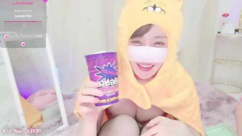 Media: A video of an Asian woman with light skin, wearing a yellow Pikachu onesie and Pikachu mask, sitting on a bed, holding a purple cup labeled \"XIBR.\