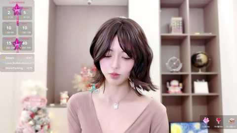 Media: Video of a young woman with shoulder-length brown hair, wearing a V-neck top and a silver necklace, standing in a modern, light-toned living room with shelves and decorative items.