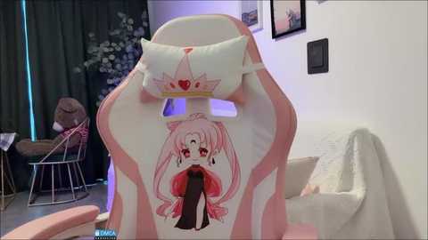 Media: Video of a pink gaming chair adorned with a cute anime girl illustration, sitting in a cozy, dimly lit room with a white couch, floral decor, and a framed picture on the wall.