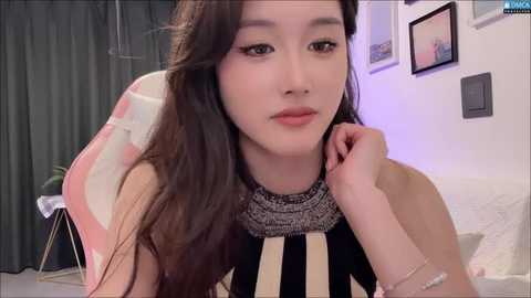 Media: Video of an Asian woman with long black hair, wearing a black and white striped top, looking contemplative, in a dimly lit bedroom with framed pictures on the wall.