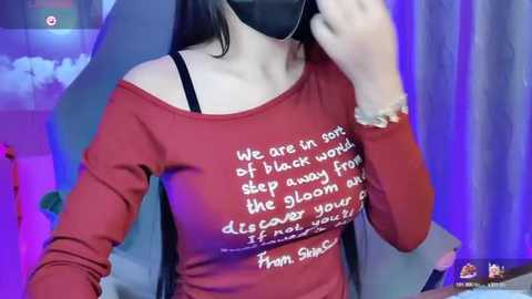 Media: A video of a woman with long, dark hair, wearing a black mask and a red off-shoulder top with a white quote on it, holding a smartphone.