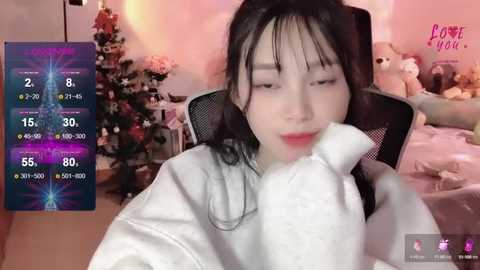 Media: Video of an Asian woman in a white bathrobe, sitting in a chair with her face in her hand. Background includes a Christmas tree, stuffed animals, and a digital overlay with health statistics.