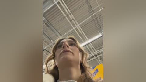 Media: A video of a woman with blonde hair, looking upwards, inside a warehouse with visible metal beams and a yellow sign.