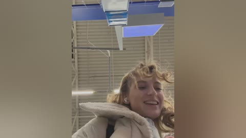 Media: A video of a woman with curly blonde hair, wearing a white jacket, laughing in a modern room with blue and white lighting fixtures and beige walls.