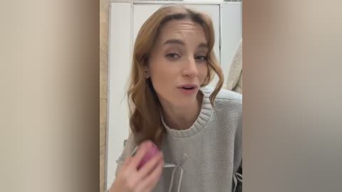 Media: A video of a young Caucasian woman with light brown hair, wearing a grey hoodie, taking a selfie in a beige-walled hallway, holding a pink object.