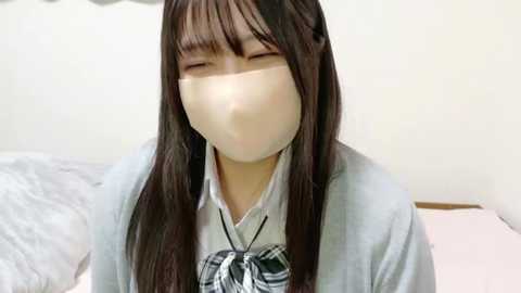 Media: A video of a young Asian woman with long dark hair, wearing a white mask, school uniform, and sitting on a bed with white sheets. The background is plain with light walls.