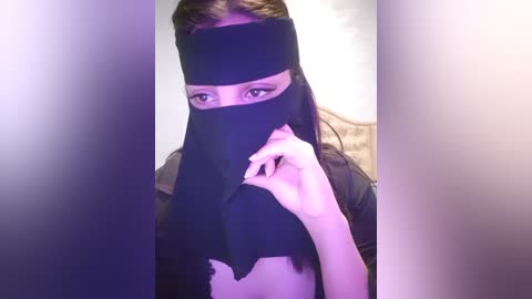 Media: Video of a young woman with light skin, dark hair, and a black face mask, holding the mask to her face, partially obscuring her eyes. Background is blurred with purple and white tones.