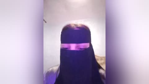 Media: Video of a person wearing a black niqab with pinkish-purple lighting, creating a surreal and slightly distorted appearance. The background is a plain white wall, adding to the mysterious and abstract atmosphere.