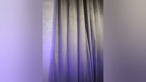 Media: A video of a vertical, full-length curtain in a muted gray color, with subtle, gradient purple lighting on the left side, creating a soft, serene ambiance. The background wall is plain and neutral.