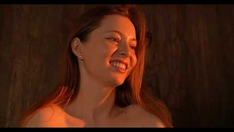 Media: Video of a smiling, fair-skinned woman with long, straight brown hair, wearing small earrings, illuminated by warm, orange light. She has a slender physique and is topless, with a dark, textured background.