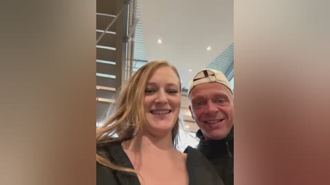 Media: A video of a smiling, fair-skinned woman with long, wavy blonde hair in a black top, standing next to a grinning, bald man in a baseball cap and black jacket. They're indoors, near a modern, glass-walled staircase.