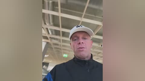 Media: Video of a Caucasian man in a black jacket and white baseball cap, standing in a modern indoor space with a ceiling of exposed beams and ductwork.