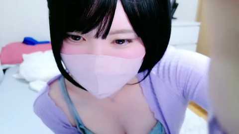 Media: A video of an Asian woman with a light complexion, short black hair, and a surgical mask, wearing a lavender cardigan over a blue bra. Background shows a messy bed with white sheets and a pink blanket.