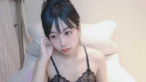 Media: A video of an East Asian woman with fair skin, black hair in a messy bun, wearing black lace lingerie, sitting on a white bed in a minimalist room with cream walls.