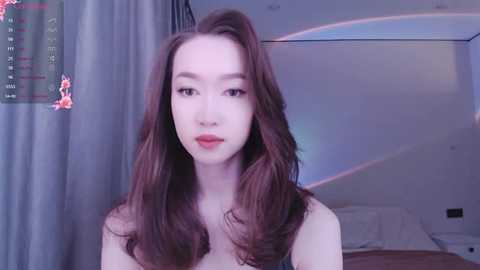 Media: Video of a young East Asian woman with long, wavy brown hair, fair skin, and red lipstick, standing in a softly lit bedroom.