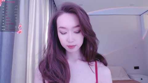 Media: Video of a young Asian woman with long, wavy brown hair, fair skin, wearing a red camisole, standing in a softly lit bedroom with a bed and window with curtains in the background.