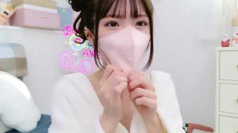 Media: Video of a young Asian woman in a white robe, wearing a pink mask, holding her hands near her face. Background features a pastel-colored room with a pink storage bin and a bed with a plaid blanket.