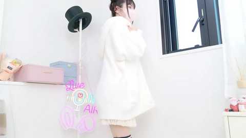 Media: Video of a young woman in a white fluffy coat, standing near a window, with a black top hat on a wall, and pastel-colored objects on a shelf in a bright, minimalistic room.