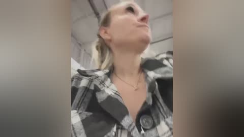 Media: A blurry video of a woman with blonde hair, wearing a black and white plaid shirt, gazing upwards. She has a necklace and is in an indoor setting with visible ceiling tiles.