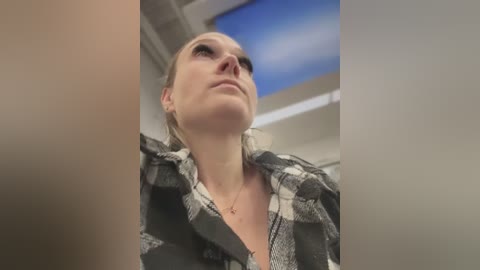 Media: Video of a blonde woman in a black and white checkered shirt, looking up with a serious expression, in a modern indoor setting with a blue sky ceiling.