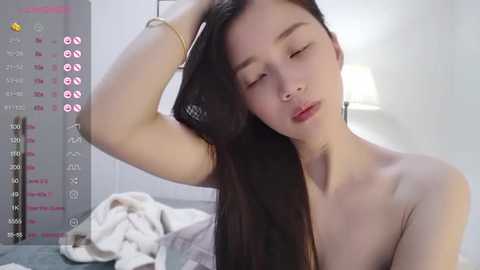 Media: Video of an East Asian woman with long black hair, light skin, and red lipstick, topless, leaning against a bed with white sheets, in a minimalist bedroom with a white lampshade.