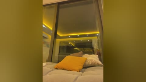 Media: Video of a modern, dimly-lit bedroom with a large bed covered in light grey sheets, featuring a mustard-yellow pillow and a white pillow. The room has a sleek design with a large window showing a cityscape at night, illuminated by yellow LED lighting.