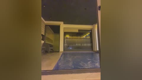 Media: Video of an indoor pool area, viewed through a doorway. The pool has a blue tiled bottom, surrounded by beige tiles. A black couch and a framed picture are visible on the left wall.