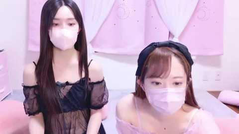 Media: Video of two young Asian women with long black hair, wearing black lace off-shoulder tops and pink face masks, in a pink-themed room with floral decor.