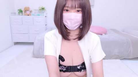 Media: Video of a young Asian woman with straight brown hair, wearing a pink surgical mask and a white cardigan, revealing a black lace bra with a floral design, in a minimalistic white bedroom with a bed and dresser.