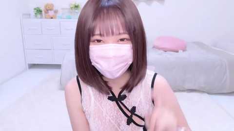 Media: Video of an East Asian woman with straight, shoulder-length brown hair, wearing a pink face mask, white lace dress, and black undergarments, standing in a minimalist bedroom with white furniture and a pink pillow.