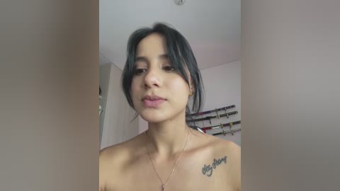 Media: A video of a young woman with medium skin tone, dark hair, and a small tattoo on her shoulder, standing in a room with a shelf of books in the background.