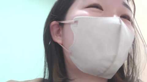 Media: Video of an Asian woman with long black hair, wearing a white surgical mask, smiling. Background is a soft, gradient blue and beige.