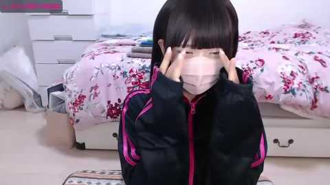 Media: A video of an Asian woman with straight black hair, wearing a black jacket with pink trim, a face mask, and kneeling in a bedroom with a bed, dresser, and floral-patterned bedding.