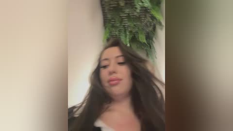 Media: Video of a young woman with long brown hair, wearing a black top, standing in front of a green fern wall hanging, her face partially blurred.