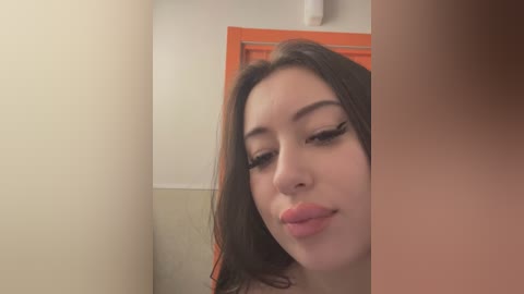 Media: A video of a young woman with fair skin, dark hair, and light makeup, puckering her lips in a bathroom with beige and orange walls.