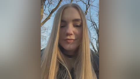 Video of a young, fair-skinned, blonde woman with long hair, wearing a black coat, smiling in a park with bare trees and a clear blue sky.