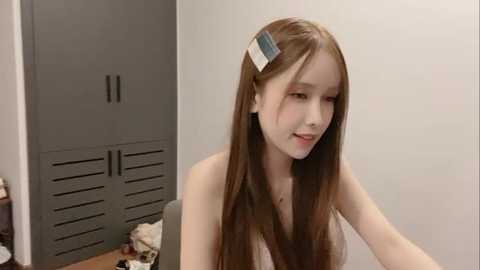 Media: Video of a young Asian woman with long, straight brown hair and a white headband, wearing a white tank top, sitting in a minimalist room with gray lockers and cluttered floor.