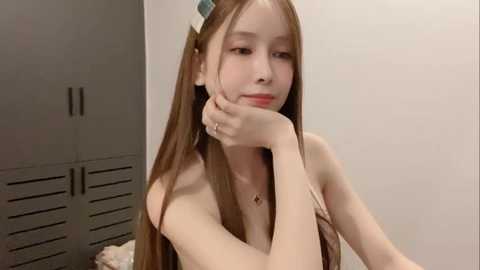 Media: Video of a young East Asian woman with long brown hair, wearing a sleeveless dress, resting her chin on her hand in a casual, introspective pose. Background shows gray lockers and a white wall.