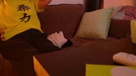 Media: A video of a person in a yellow T-shirt, black pants, and a white sock, sitting on a brown couch with green and white pillows, holding a white remote.