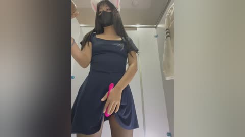 Media: Video of a woman in a dark blue dress with a pink vibrator, black mask, and bunny ears, standing in a beige-tiled bathroom.