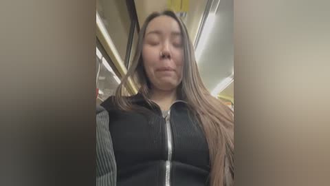 Media: A video of an Asian woman with long, straight brown hair, wearing a black and white striped shirt, eyes closed, standing in a dimly lit subway car.