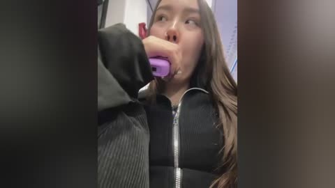 Media: Video of a young woman with long brown hair, wearing a black ribbed sweater, using a purple toothbrush. She is in a bathroom with a white sink and mirror in the background.