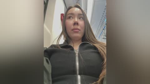 Media: Video of a young woman with long brown hair, wearing a black jacket, looking upwards in a subway train. The background shows metallic train compartments and a red emergency button.