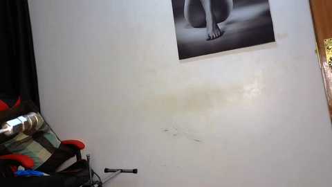 Media: Video of a white wall with a black and white framed video of a nude person, partially covered by a yellow stain. Below the wall, a cluttered table holds a can, a red and black helmet, and a green bottle.