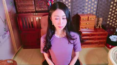 Media: Video of an Asian woman with long black hair, wearing a purple shirt, sitting on a tatami mat in a traditional Japanese room with wooden furniture and decorative items.